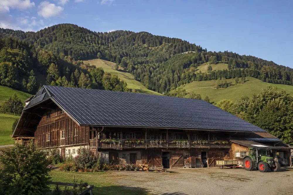Große Holzscheune, das komplette Dach mit dunklen Solarmodulen belegt, dahinter bewaldete Hänge und ein Traktor im Hof